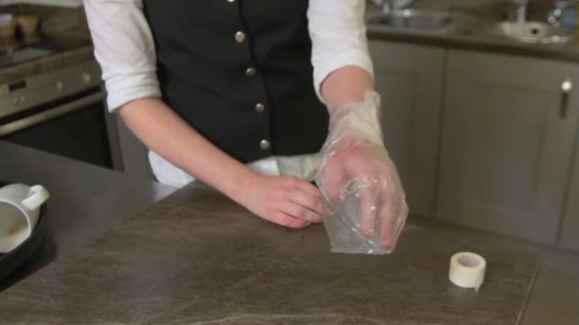 A Person Is Wearing a Plastic Bag Over Their Hand in A Kitchen, Likely Treating a Burn, with A Roll of Medical Tape on The Counter Nearby